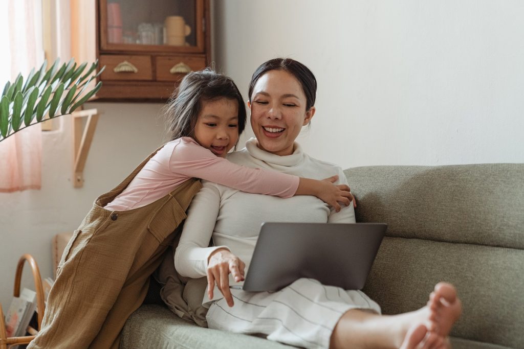 Mom sitting on couch looking at laptop while child hugs her.