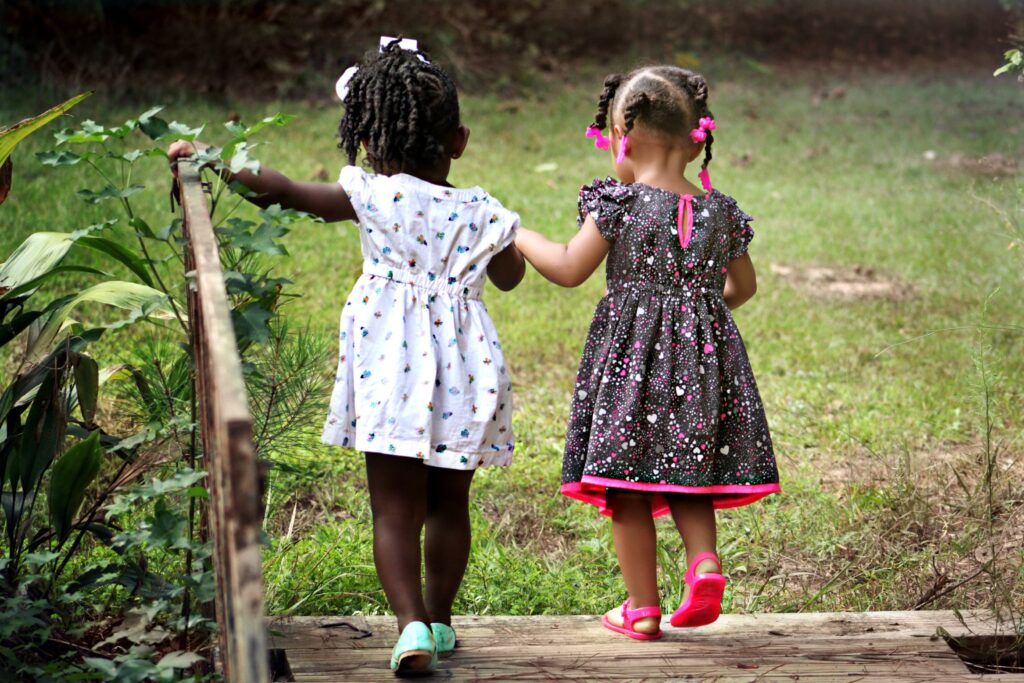 Two young girls walk hand in hand on a wooden path in a garden. They wear colorful dresses and sandals, conveying a sense of friendship and adventure.