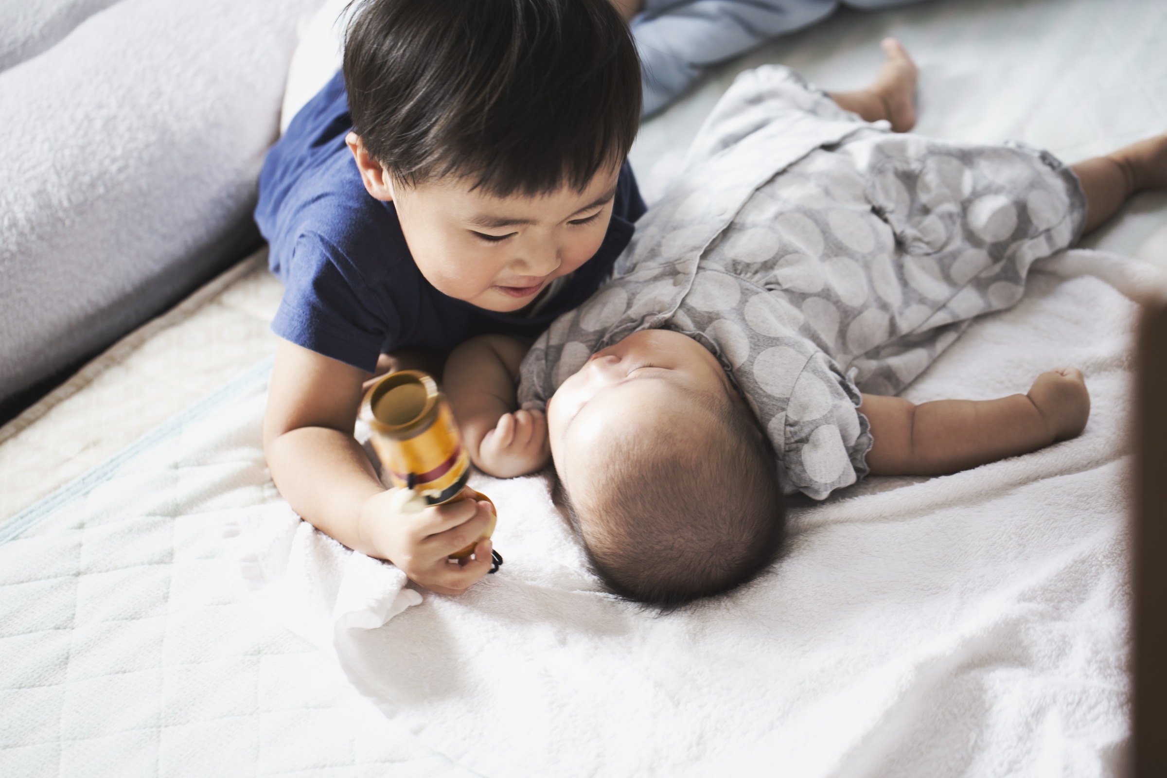 A toddler boy holding a toy and smiling at his baby sibling. 