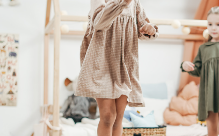 A young girl in a beige dress joyfully jumps in a cozy, playful bedroom. A wicker basket and colorful pillows add warmth to the cheerful scene.