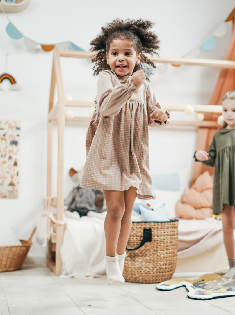 A young girl in a beige dress joyfully jumps in a cozy, playful bedroom. A wicker basket and colorful pillows add warmth to the cheerful scene.