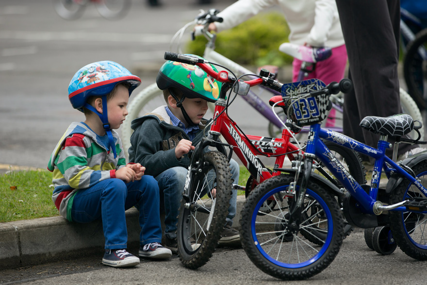 Two young boys wearing helmets sit on a curb, intently examining bicycles. Nearby are colorful bikes and other children, suggesting a playful, curious atmosphere.