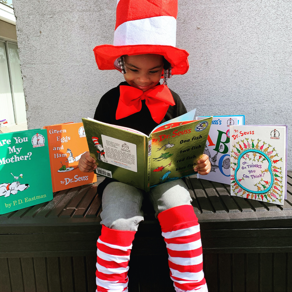 A young child, dressed as the Cat in the Hat with a red and white hat and bow, reads "One Fish, Two Fish" by Dr. Seuss. Other Dr. Seuss books are displayed nearby. The scene is colorful and playful.