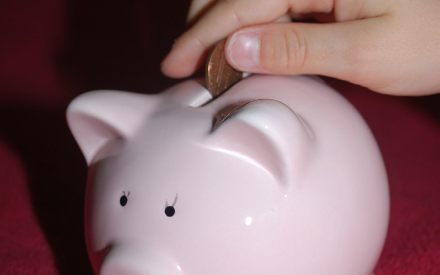 A hand places a coin into a cute pink ceramic piggy bank. The background is a soft red fabric, creating a cozy and nurturing atmosphere.