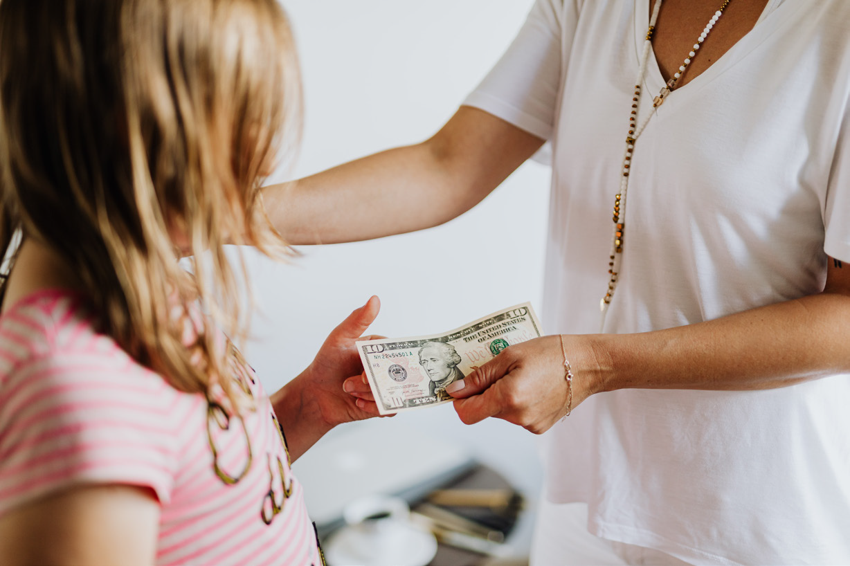A woman in a white shirt hands a ten-dollar bill to a child in a pink striped top. The scene conveys a feeling of trust and care..