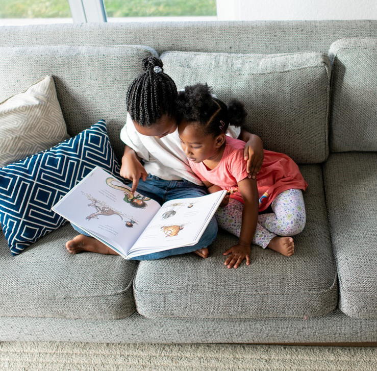 Two children sit closely on a gray sofa, reading a picture book together. The child on the left wears a white shirt, the one on the right, a pink dress.