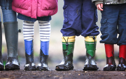 Children wearing muddy rain boots in various colors—black, blue, green, and red—stand on a dirt path, suggesting playful outdoor activity.