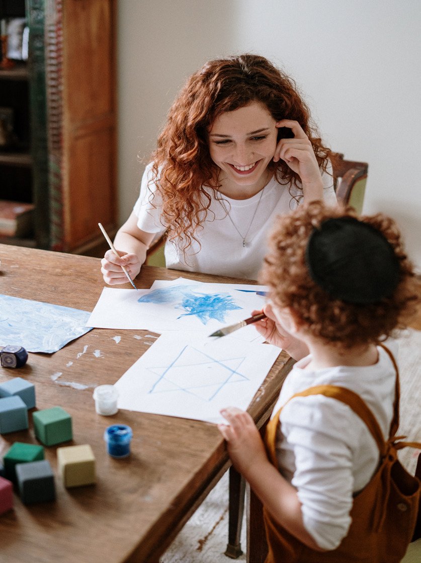 A smiling woman and a child with curly hair paint together at a wooden table. Art supplies and colorful blocks are scattered around, creating a creative and warm atmosphere.