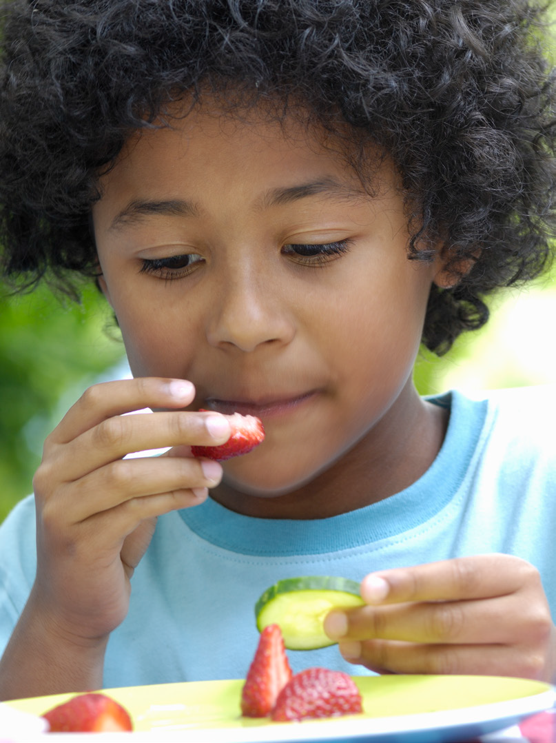 Young child with curly hair, wearing a light blue shirt, holds a slice of cucumber and a strawberry. They appear thoughtful and focused. Background is lush and green.