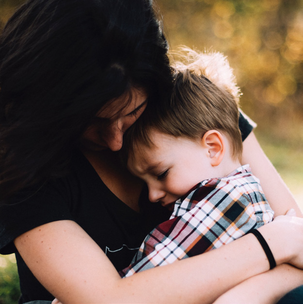 A woman embraces a young boy wearing a plaid shirt, both showing affection and comfort. They are outdoors with a blurred, warm-toned background.