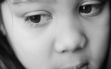 Close-up black-and-white portrait of a young child with long hair, looking downwards pensively. The expression conveys a sense of contemplation.