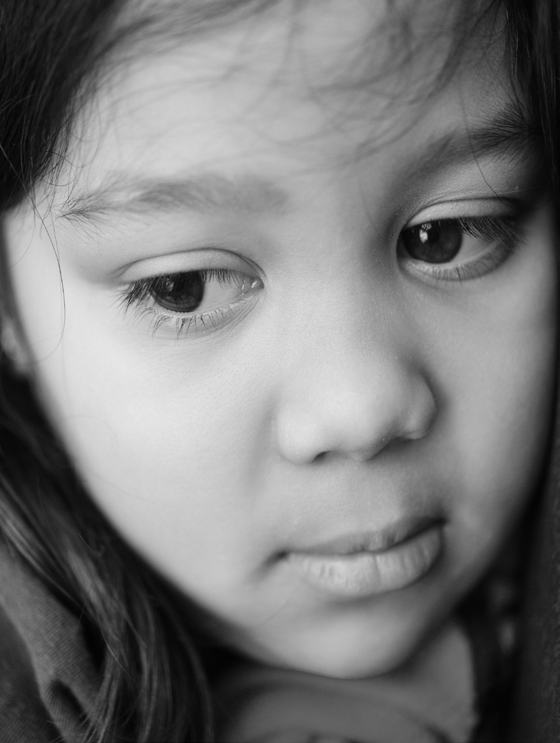 Close-up black-and-white portrait of a young child with long hair, looking downwards pensively. The expression conveys a sense of contemplation.