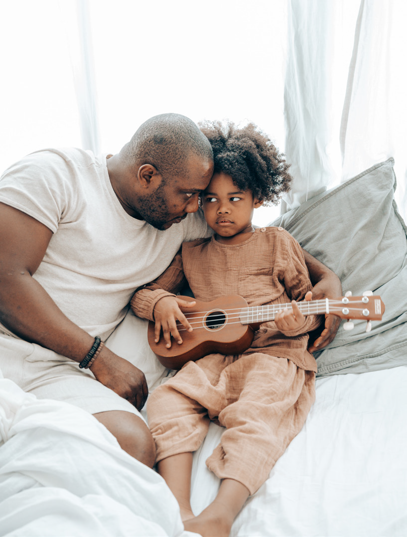 Father and child sit on a bed with soft lighting, sharing a tender moment. The child holds a small guitar, wearing a focused expression, conveying warmth and connection.