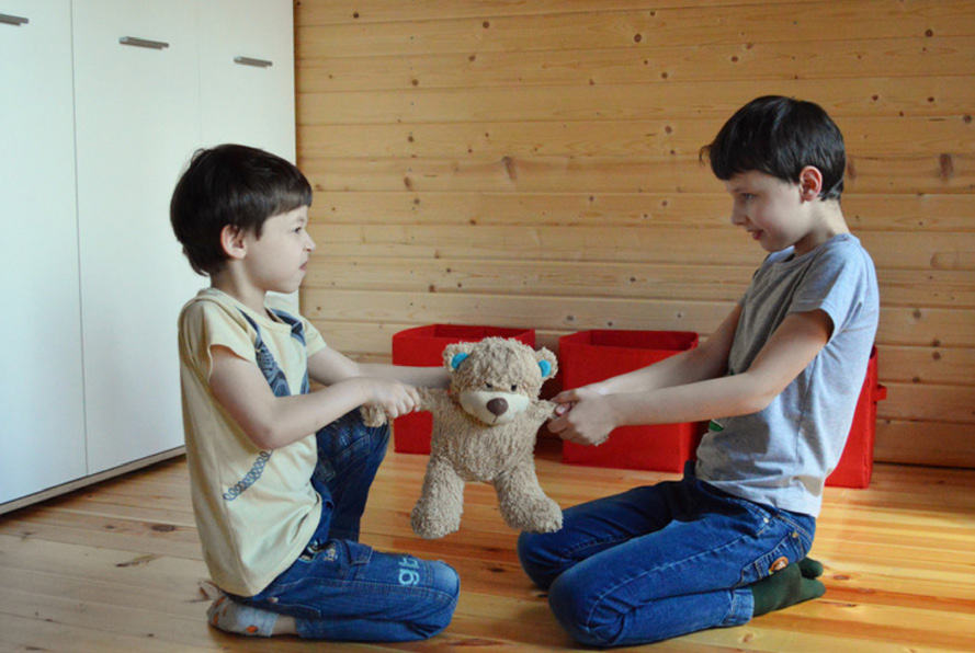 Two children sit on a wooden floor, both tugging at a teddy bear with colorful ears