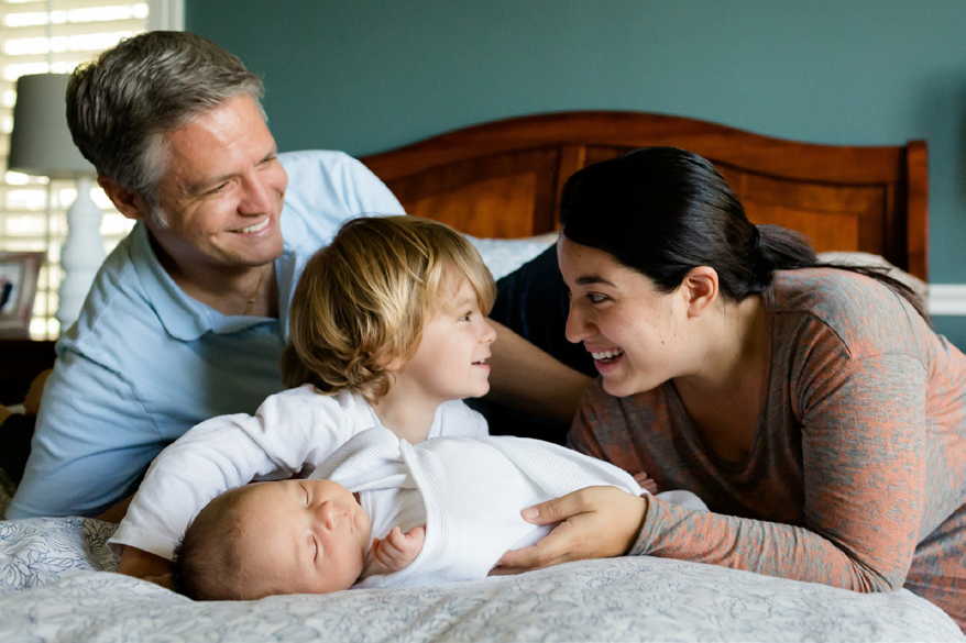 A joyful family, including a father, mother, and toddler, smiles at a sleeping newborn wrapped in a white blanket on a bed, conveying warmth and happiness.