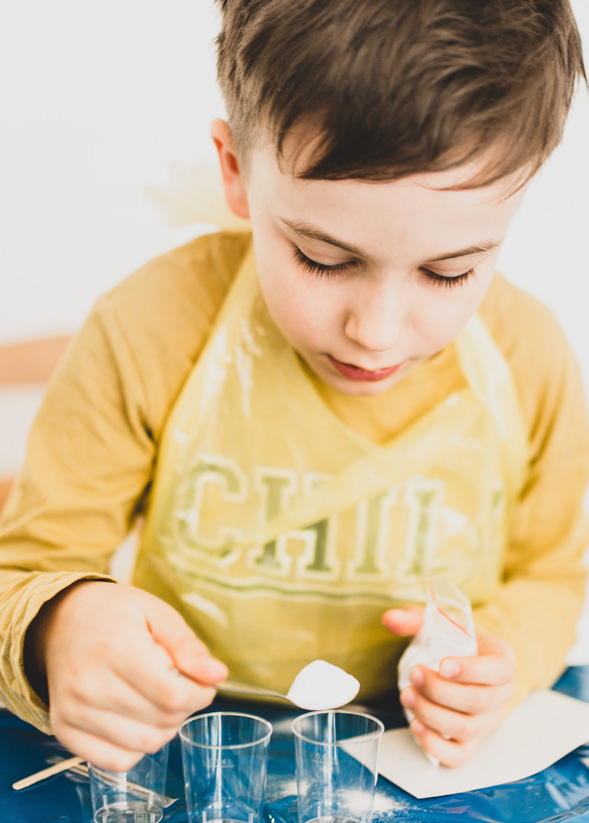 A young boy in a yellow shirt and apron focuses on measuring white powder into clear cups with a spoon