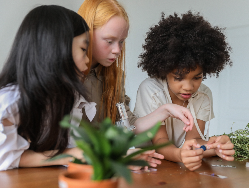 Three children engaged in a science activity. They are focused on using a microscope and examining small objects. A plant sits on the table, creating a learning atmosphere.