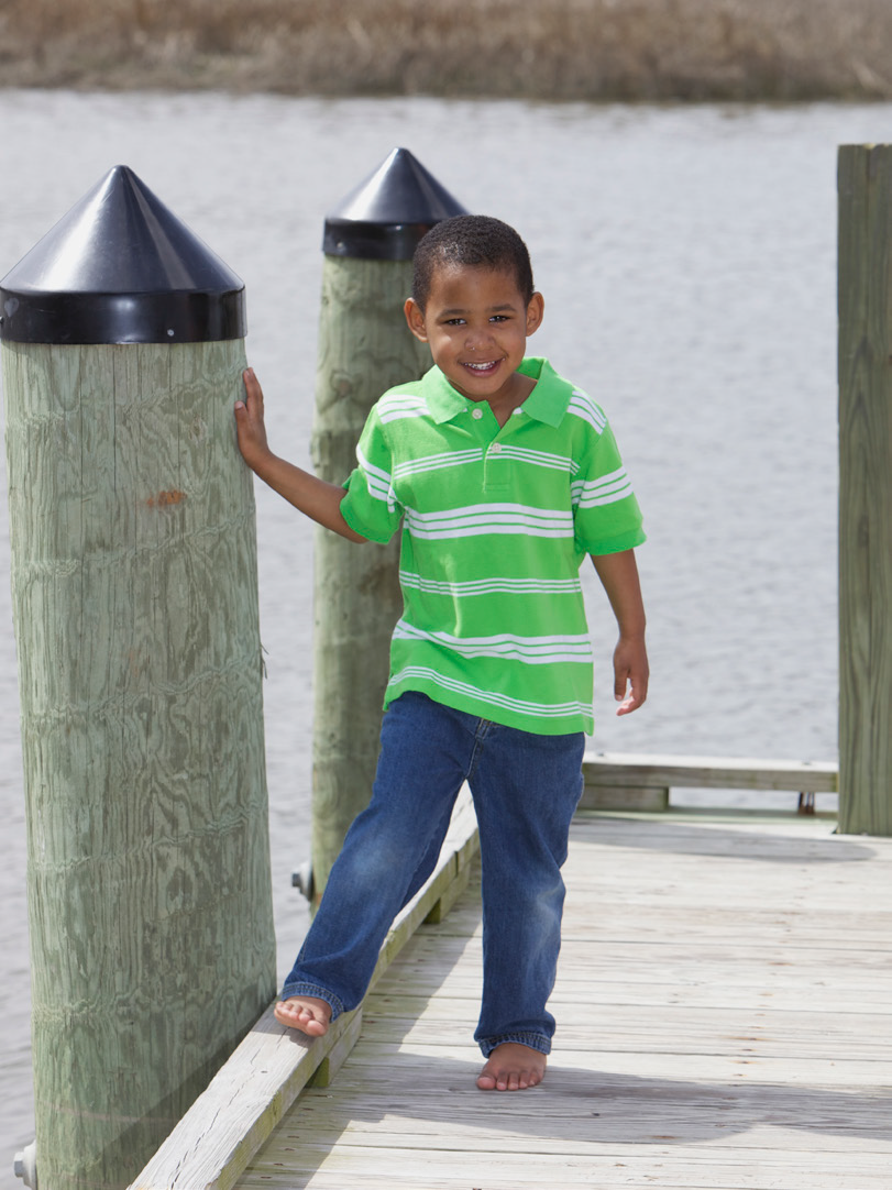 Young boy in a bright green striped shirt and jeans smiles while standing on a wooden dock, touching a post. The background shows calm water.