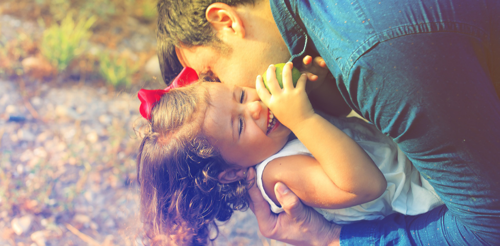 A man in a blue shirt lovingly hugs a laughing child with curly hair and a red bow outdoors.