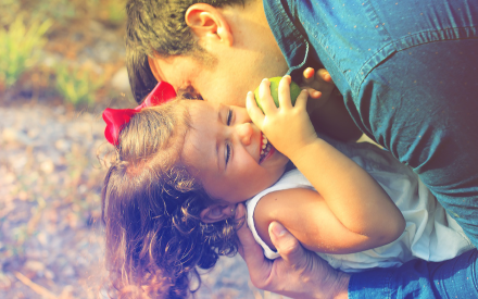 A man in a blue shirt lovingly hugs a laughing child with curly hair and a red bow outdoors.