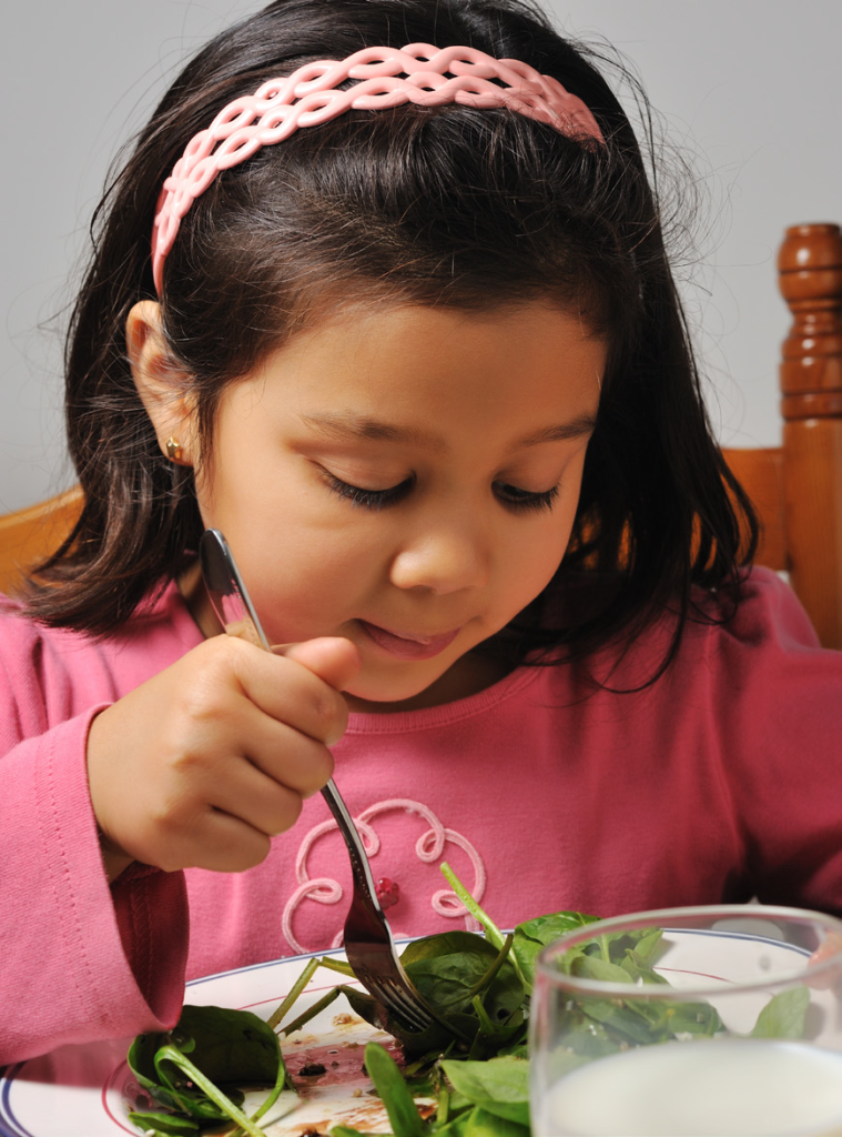 A young girl in a pink top and headband is focused on eating a salad with a fork. A glass of milk is nearby, creating a calm, healthy scene.