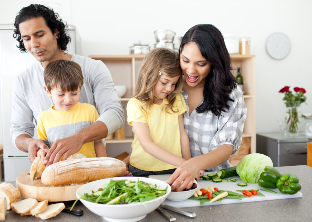 A family of four enjoys cooking together in a kitchen. The father slices bread, the mother and daughter smile while preparing vegetables, and the son watches. Veggies and a salad bowl are on the counter, creating a joyful and collaborative atmosphere.