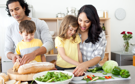 A family of four enjoys cooking together in a kitchen. The father slices bread, the mother and daughter smile while preparing vegetables, and the son watches. Veggies and a salad bowl are on the counter, creating a joyful and collaborative atmosphere.