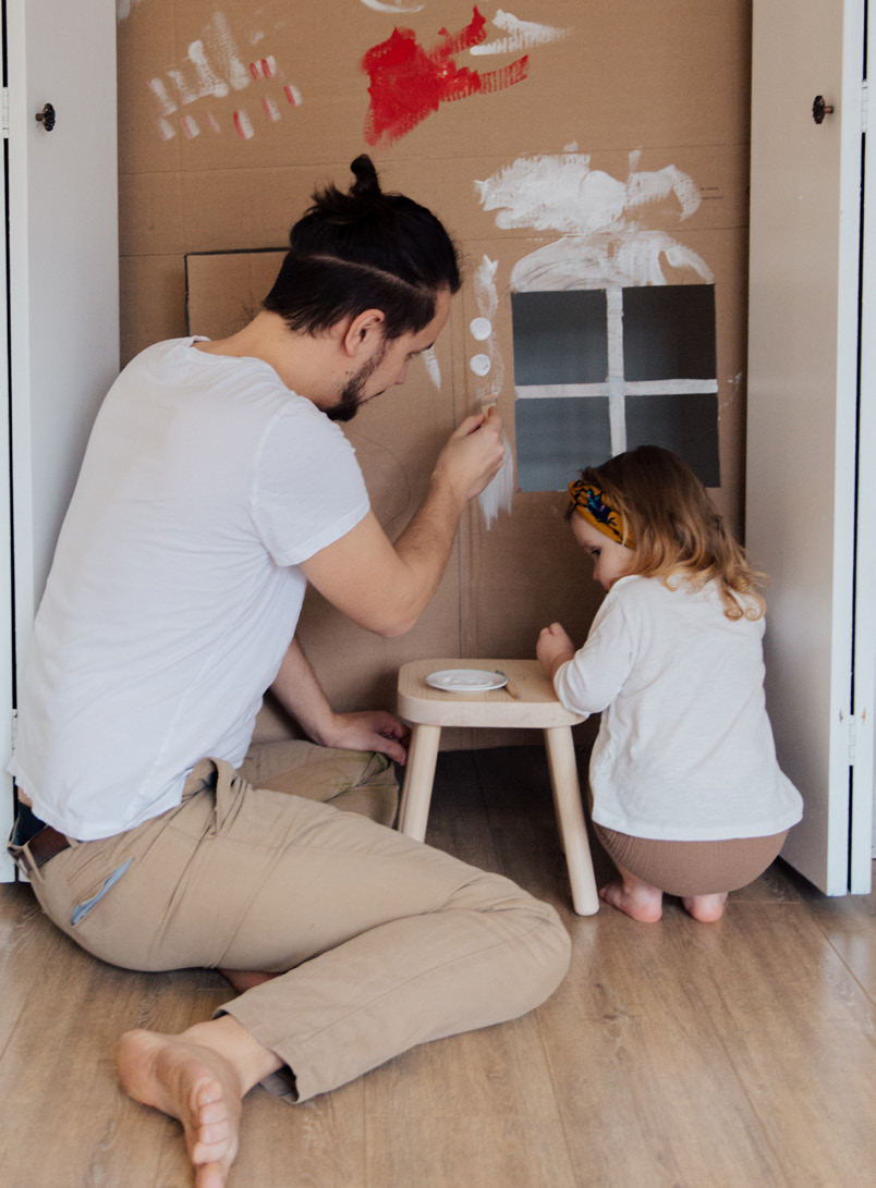 A man and a young child are painting on a large cardboard box. The man wears a white shirt and beige pants, while the child wears a headband. They appear focused and engaged in the activity.