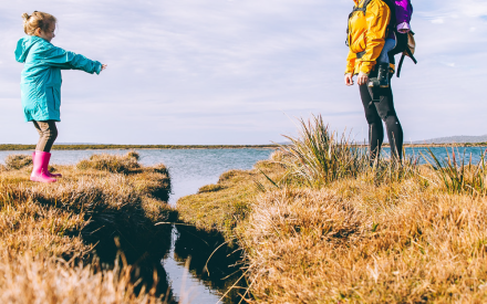 A woman in an orange jacket and child in a blue coat with pink boots face each other across a stream in a grassy, coastal landscape under a blue sky.