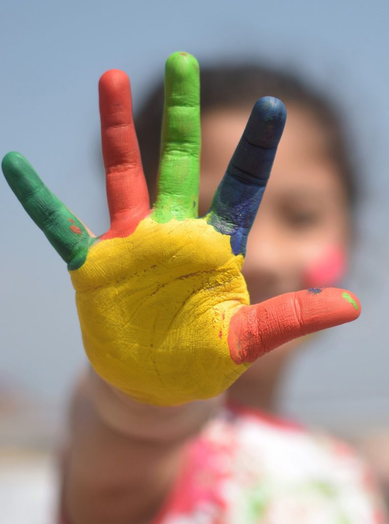 A child's hand covered in bright paint—red, green, yellow, orange, and blue—extends toward the camera against a blurred background, conveying creativity and playfulness.