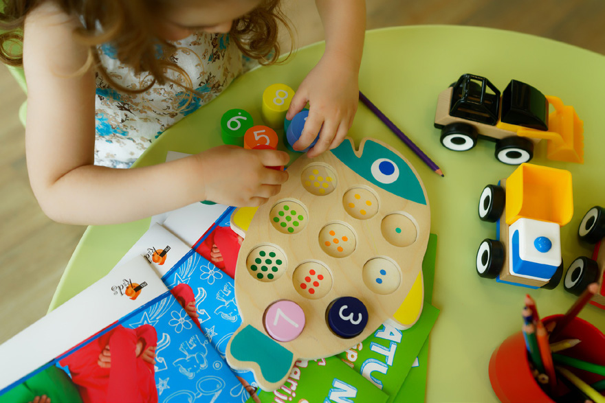 A child plays with a colorful wooden fish puzzle on a round table. Toy trucks, books, and colored pencils are scattered, conveying a playful, learning atmosphere.