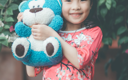 Smiling girl in a floral dress hugging a blue teddy bear, standing outside with green foliage and pink flowers in the background, exuding joy and warmth.