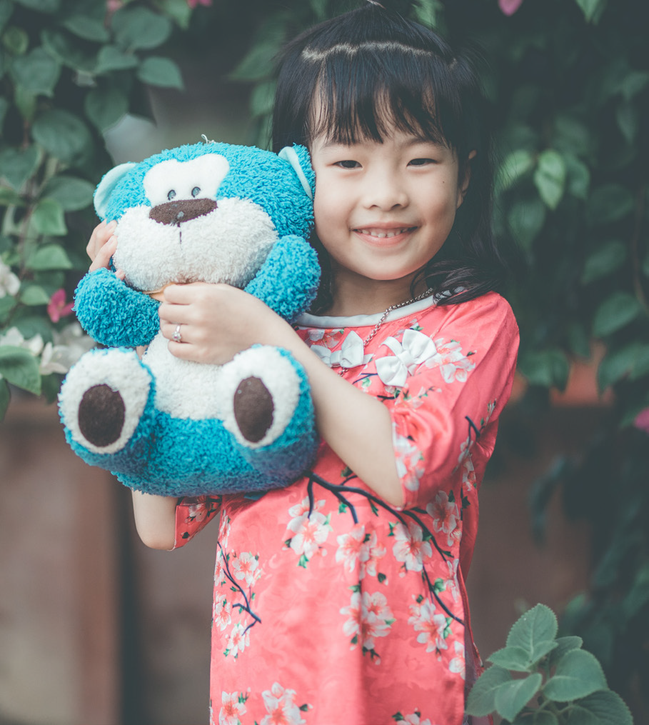 Smiling girl in a floral dress hugging a blue teddy bear, standing outside with green foliage and pink flowers in the background, exuding joy and warmth.