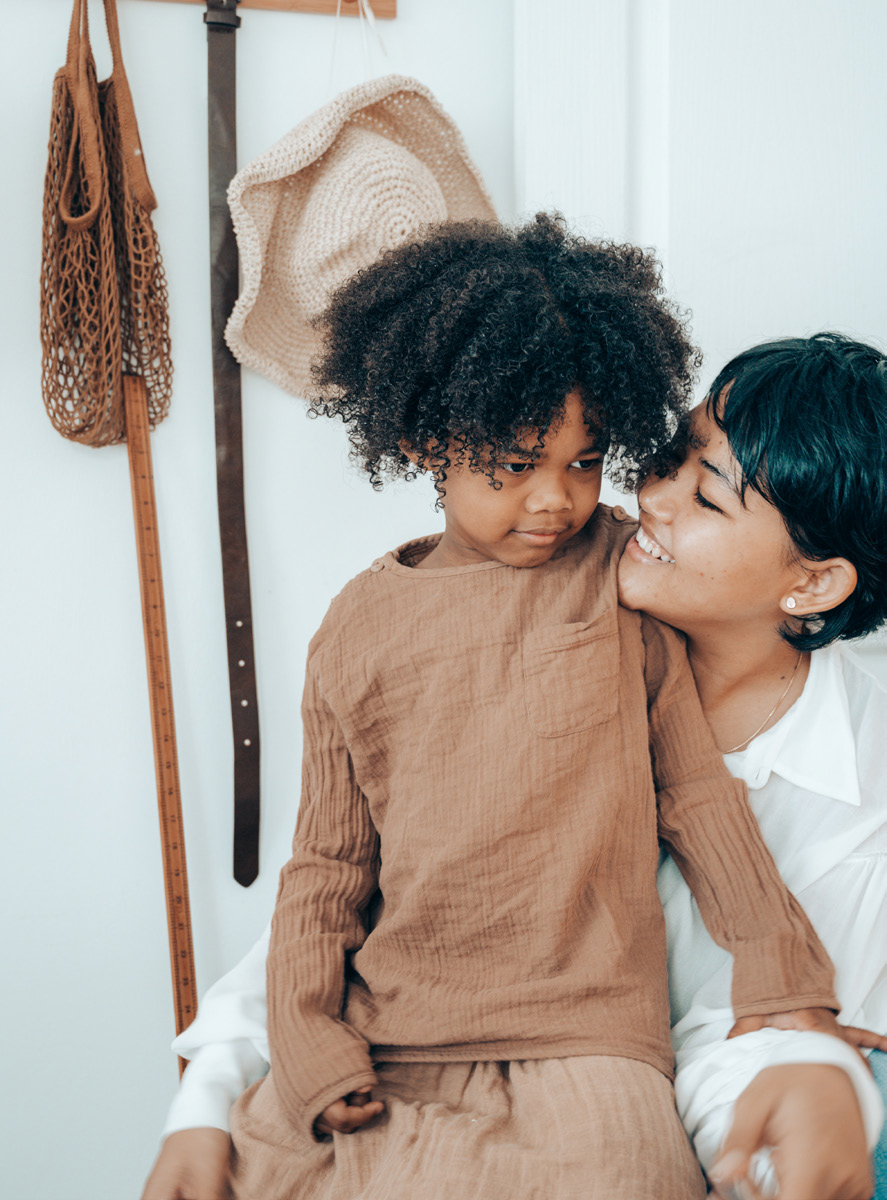 A smiling adult embraces a child dressed in brown. The child looks thoughtful. Behind them, a wall displays a woven bag, a hat, and a hanging belt. Warm, cozy atmosphere.