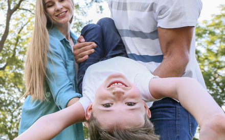 A joyful family scene in a park. A man and woman hold a laughing child upside down. Sunlight filters through trees, highlighting their smiling faces.