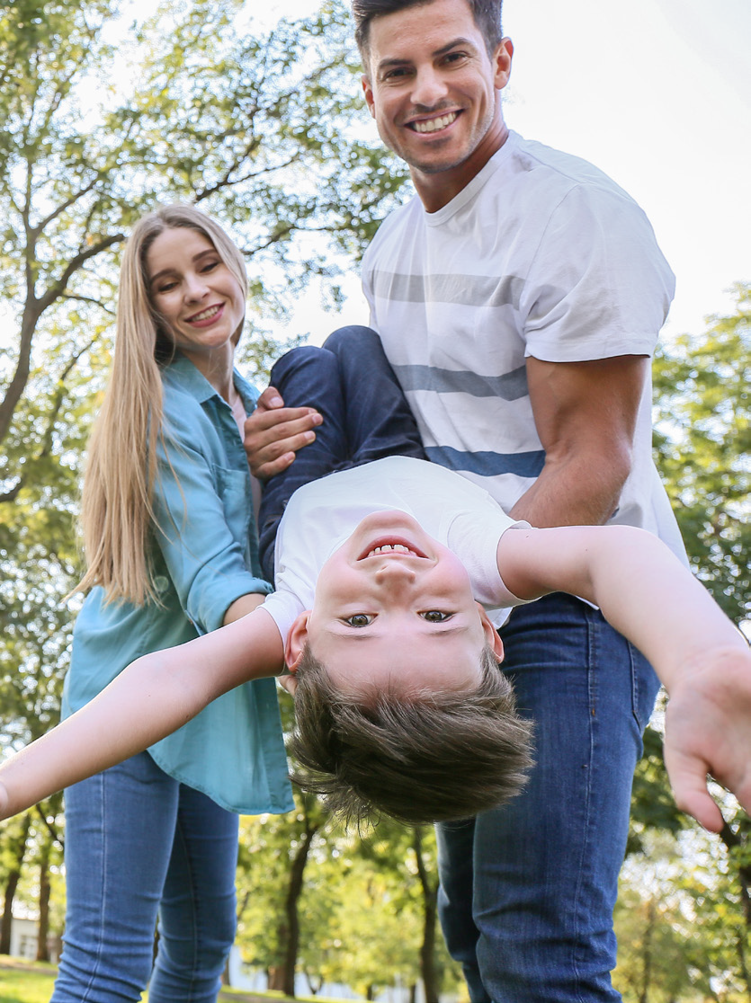 A joyful family scene in a park. A man and woman hold a laughing child upside down. Sunlight filters through trees, highlighting their smiling faces.