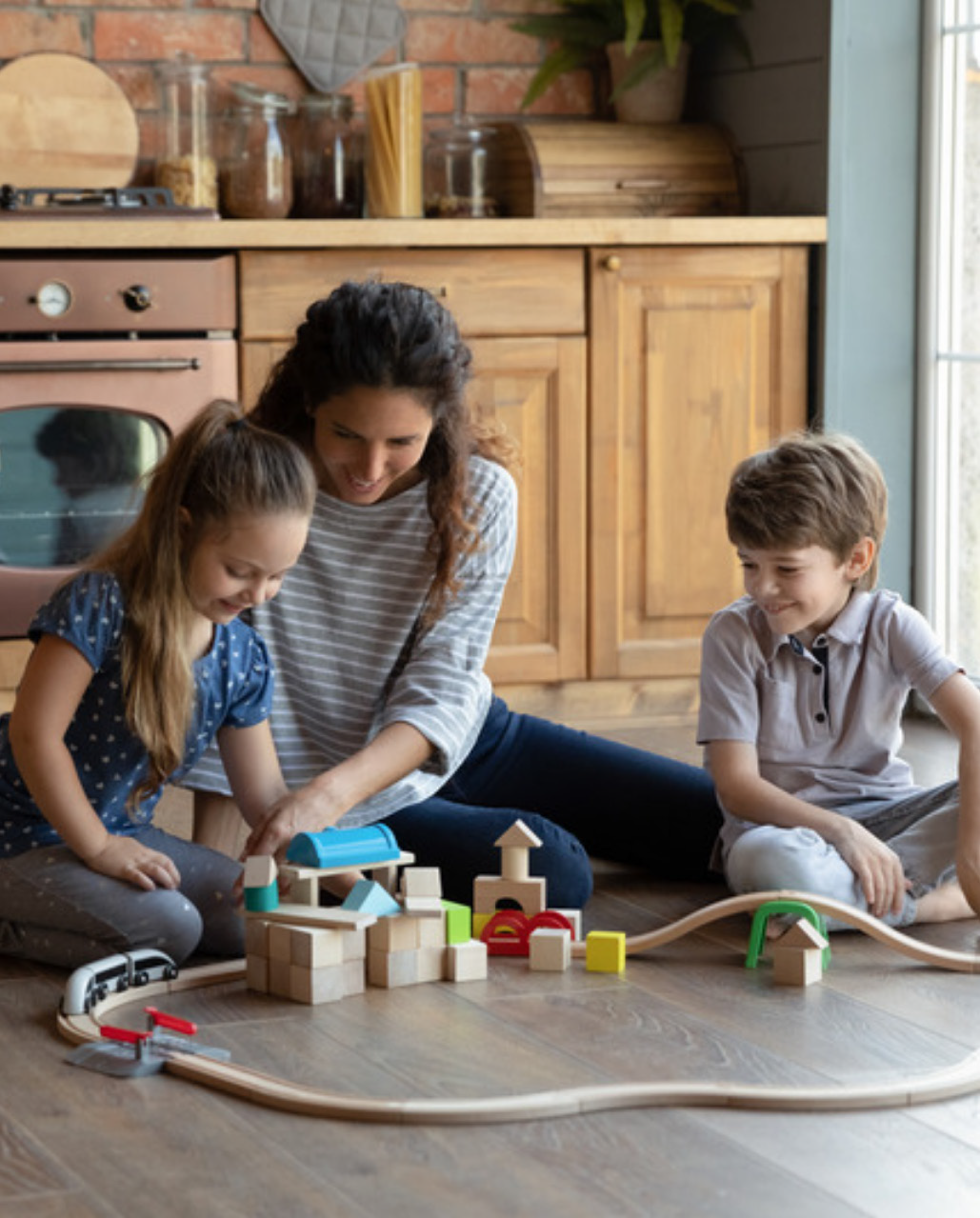 A woman and two children joyfully play with wooden blocks forming a train set in a cozy kitchen. The atmosphere is warm and engaging.