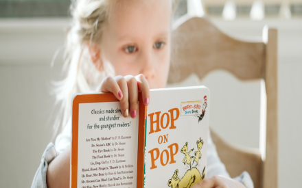 A young girl with braided hair intently reads "Hop on Pop" by Dr. Seuss. She is seated at a wooden table in a sunlit room, conveying focus and curiosity.