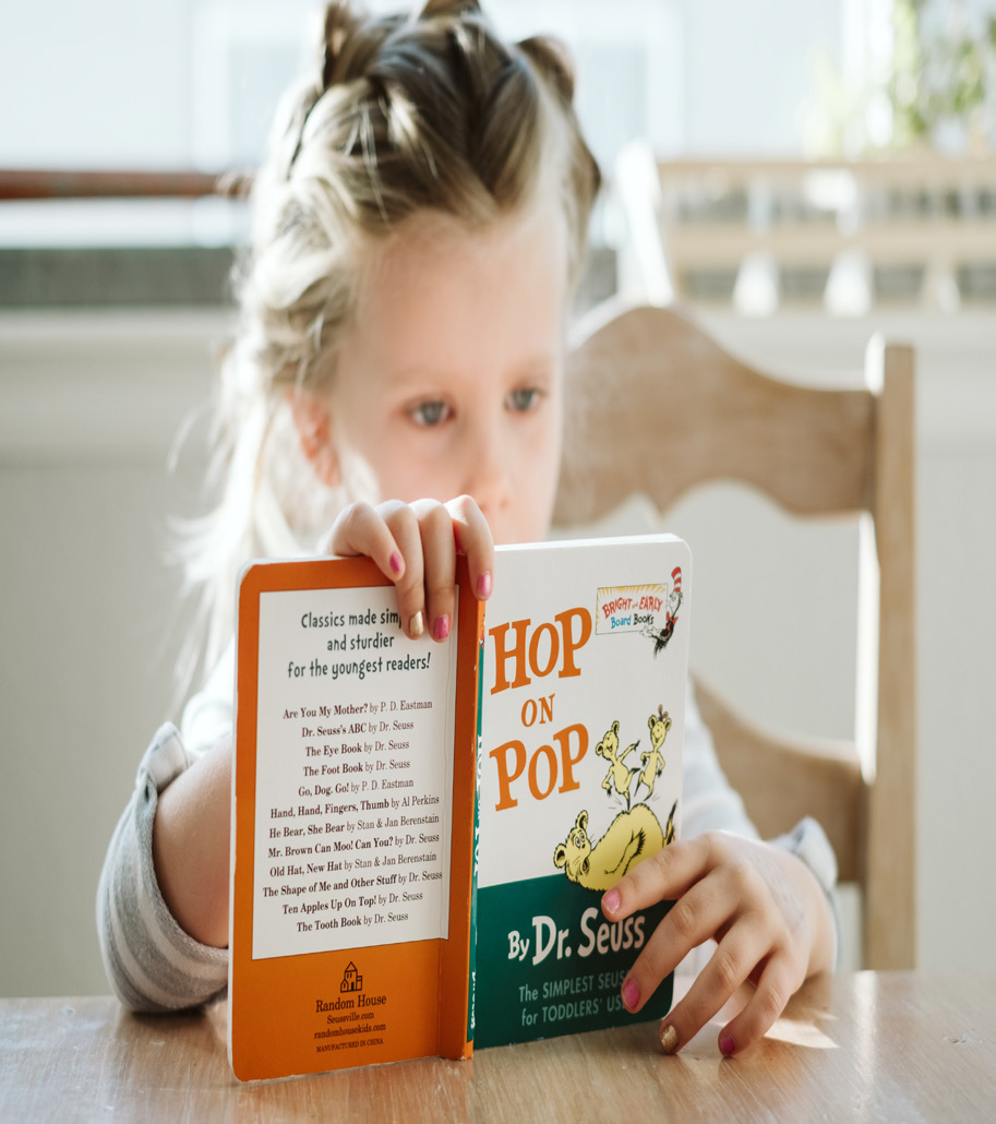 A young girl with braided hair intently reads "Hop on Pop" by Dr. Seuss. She is seated at a wooden table in a sunlit room, conveying focus and curiosity.