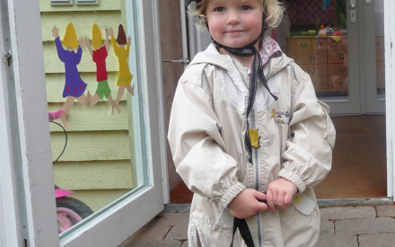 A smiling child wearing a helmet and beige jacket stands holding a blue bag outside a bright, inviting doorway. Colorful cutouts decorate the glass.