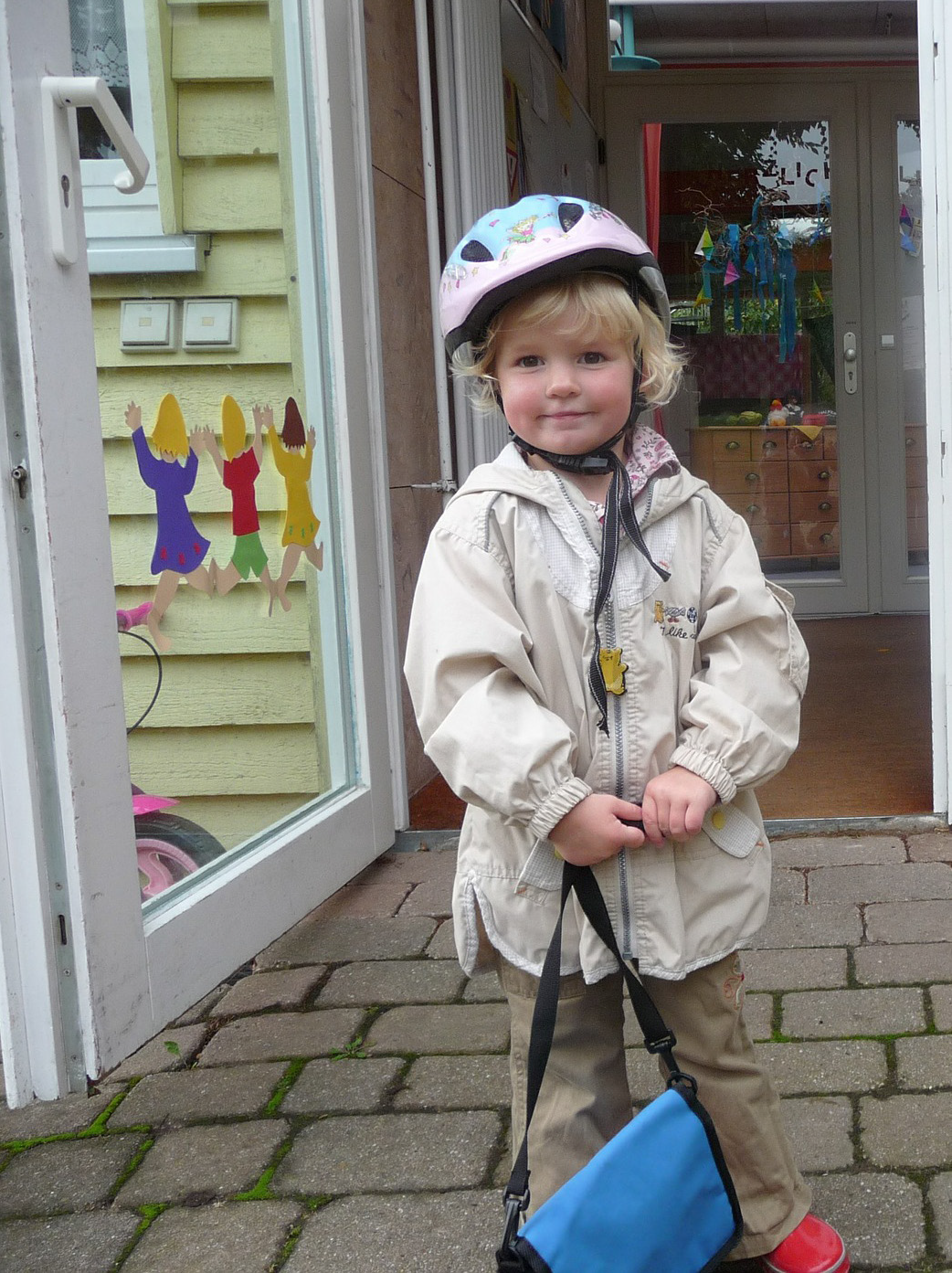A smiling child wearing a helmet and beige jacket stands holding a blue bag outside a bright, inviting doorway. Colorful cutouts decorate the glass.