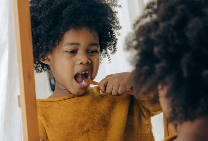 A young child in a yellow shirt brushes their teeth, looking into a mirror. The scene is bright and focused on healthy morning habits.