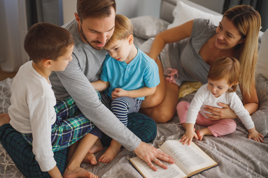 A family of five sits on a bed in pajamas, reading a book together. The scene is cozy and intimate, with warm smiles and a relaxed atmosphere.