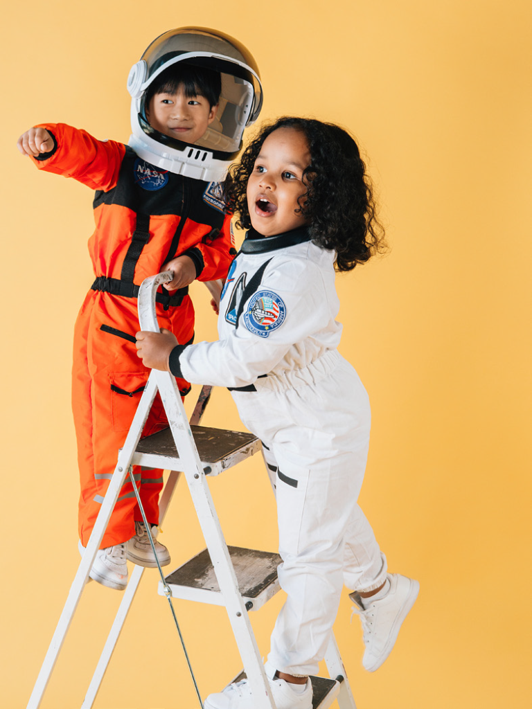 Two children in astronaut costumes are on a ladder against a yellow background. One wears a helmet and points forward, both smiling energetically. Space-themed play.