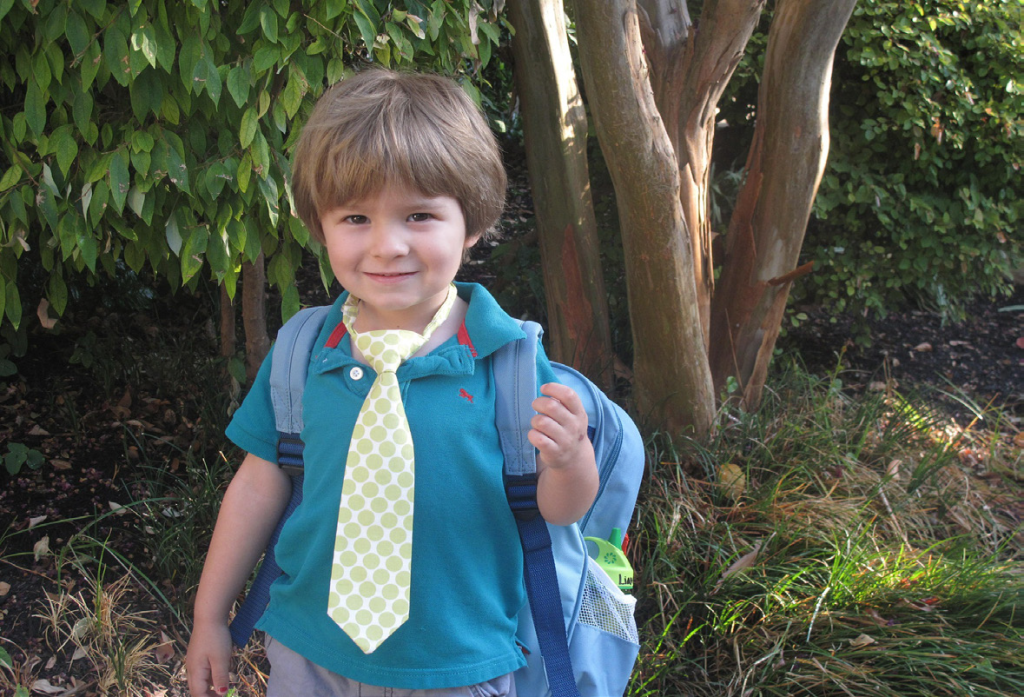 A young child stands outdoors, smiling, wearing a teal shirt, lime polka dot tie, and backpack. Surrounded by green foliage, conveying a cheerful, ready-for-school vibe.