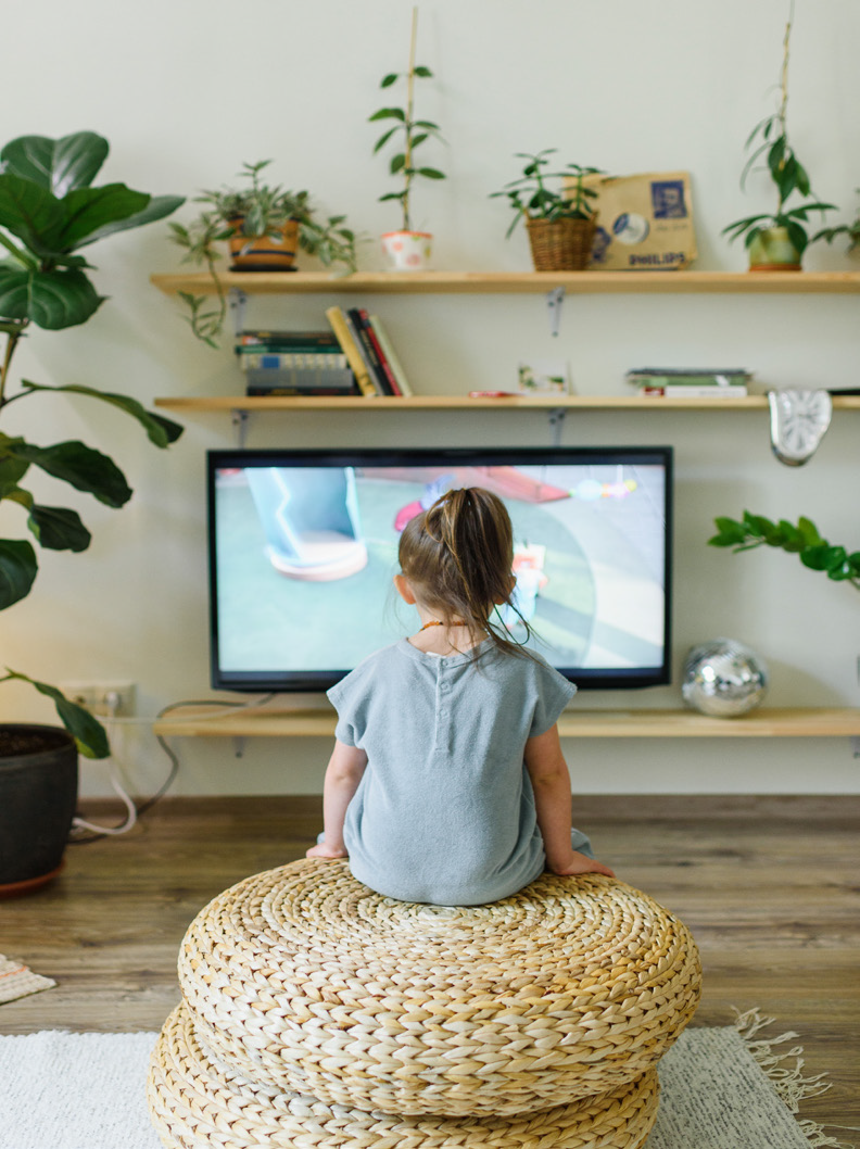 A young child sits on stacked woven cushions, watching a TV screen displaying colorful animation. The room has wooden shelves with plants and books, creating a cozy atmosphere.