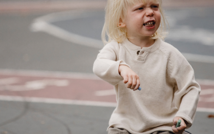 A young child with blonde hair kneels on a chalk-drawn pavement, holding chalk. The child wears a beige sweater and looks irritated.