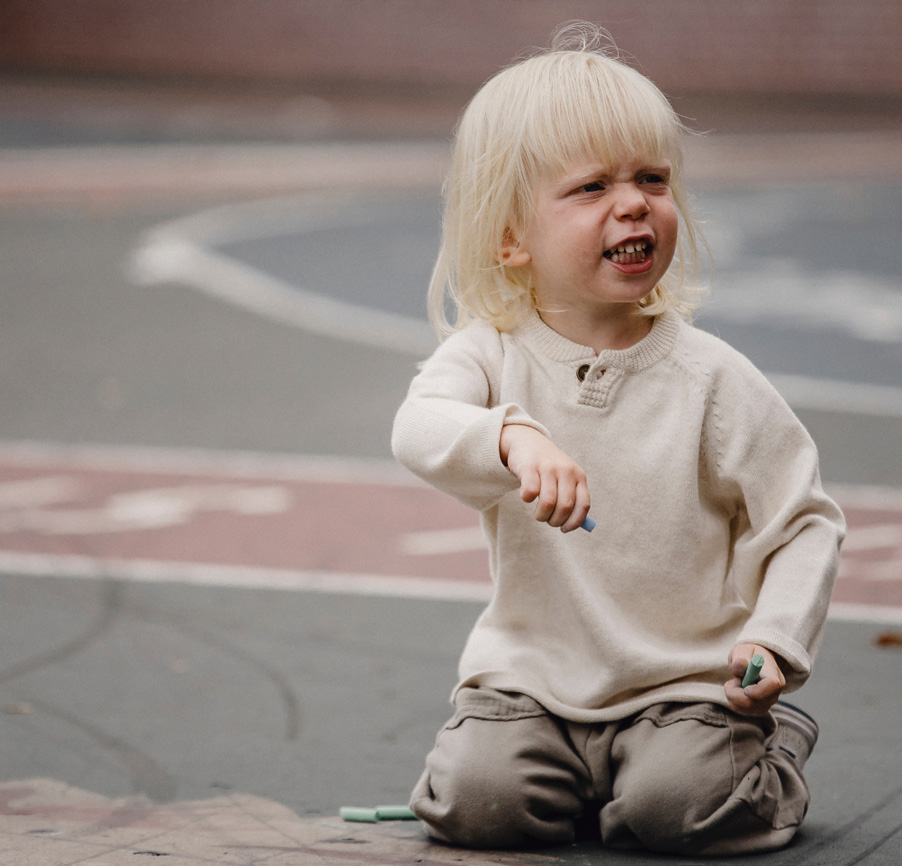 A young child with blonde hair kneels on a chalk-drawn pavement, holding chalk. The child wears a beige sweater and looks irritated.