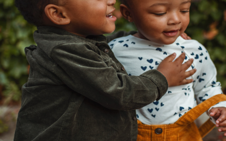 Two toddlers stand close together outdoors, smiling. One wears a green jacket, and the other sports a heart-patterned shirt and yellow corduroy skirt. Warm, playful mood.