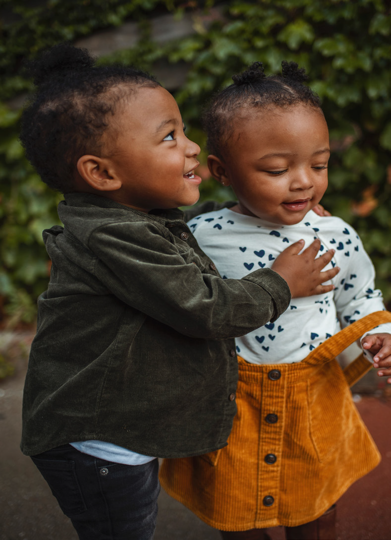 Two toddlers stand close together outdoors, smiling. One wears a green jacket, and the other sports a heart-patterned shirt and yellow corduroy skirt. Warm, playful mood.